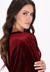Burgundi bársonydressz hosszú ujjal, hátsó kivágással és puha textúrával. Áramló kialakítása és testhez álló szabása kiemeli.