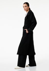 Abrigo negro largo tipo trench con cintura ceñida por un cinturón, grandes solapas y bolsillos laterales, combinado con pantalones negros y zapatillas blancas. Textura suave.