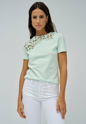 Camiseta de algodón verde menta con detalle de encaje floral en los hombros. Combinada con jeans blancos ajustados, que muestran un look casual pero refinado.