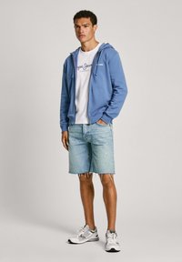 Zila rāvējslēdzēja kapuce virs baltas T-krekla ar logo, apvienota ar gaišiem džinsu šortiem un baltām kedām. Iekļauj priekšējo kabatu un auklas.