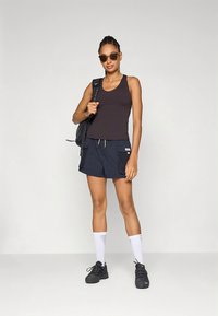 Sort tanktop, marineblå cargobukser med lommer, hvide sokker og sorte sneakers. Tilbehør inkluderer solbriller og en sort rygsæk.