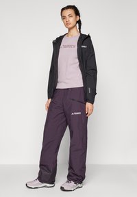 Sort vandtæt jakke, lyse lilla T-shirt med "TERREX" logo, mørklilla bukser med lynlåse, og lyse lilla sko.