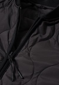 Giacca trapuntata nera con chiusura a zip, caratterizzata da una texture liscia, collo a coste e motivo di cuciture arrotondate. Tiretti sulla cerniera.