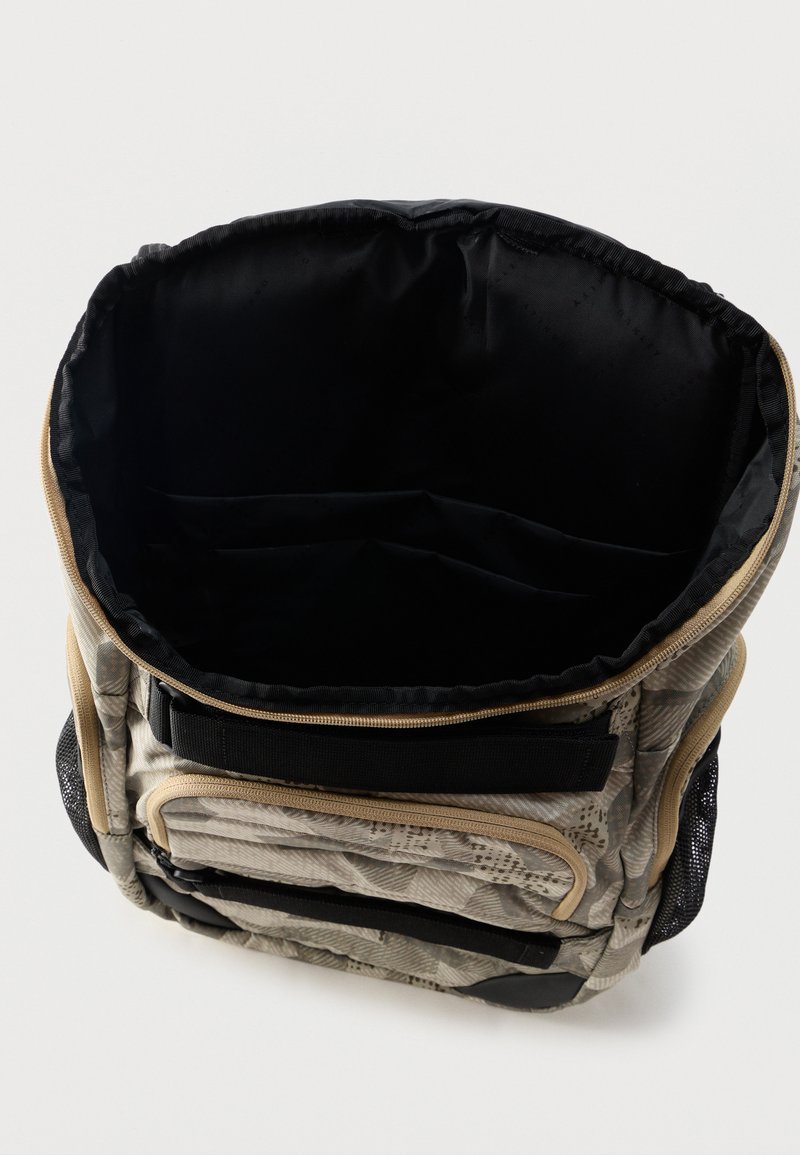 Sac à dos beige et noir à motifs avec compartiment principal ouvert, poche avant zippée et poches latérales en filet sur fond blanc.