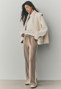 Gilet en polaire crème superposé à un sweat-shirt blanc, associé à un pantalon de survêtement beige avec des rayures sur les côtés et des chaussures légères.