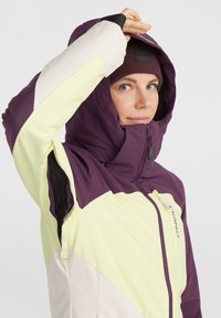 Lila und gelbe Skijacke mit hohem Kragen, Reißverschluss und verstellbarem Kapuze. Mit Kontrastpaneelen und belüfteten Einsätzen aus Mesh.