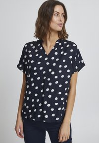 Granatowa bluzka w białe kropki, krótki rękaw i dekolt w serek. Posiada subtelną teksturę oraz marszczenia przy dekolcie.