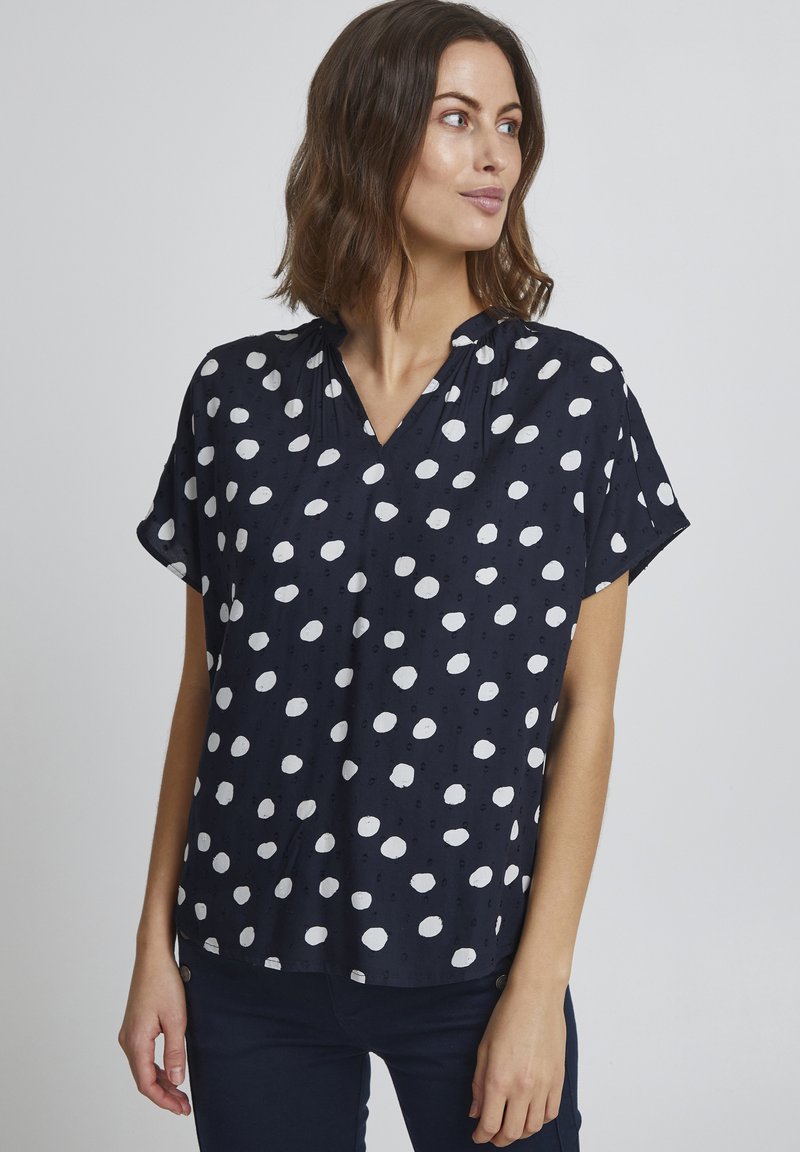 Granatowa bluzka w białe kropki, krótki rękaw i dekolt w serek. Posiada subtelną teksturę oraz marszczenia przy dekolcie.