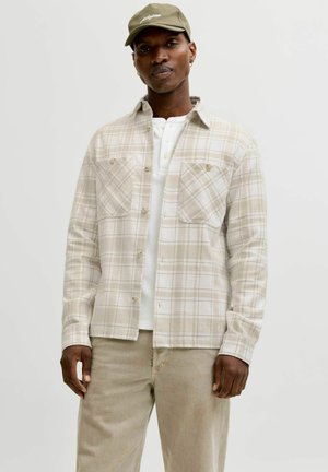 Mand iført beige ternet skjorte med knapper over en hvid henley, beige bukser og olivengrøn kasket, stående mod en ensfarvet lys baggrund.