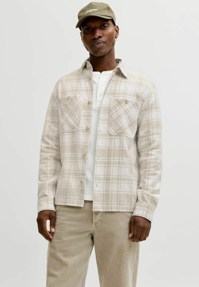 Mann iført beige rutete skjorte med knapper over hvit henley, beige bukser og olivengrønn caps, stående mot en ensfarget lys bakgrunn.