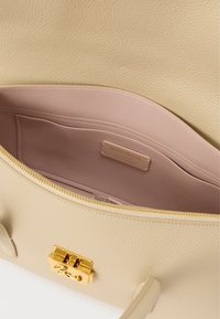 Beige gestructureerde leren handtas met gouden sluiting, deels open om de gladde roze binnenzak te tonen met het label "Coccinelle Authentic Italian Leather."
