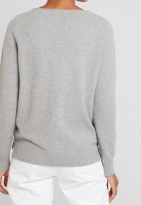 Szary dziergany sweter o luźnym kroju, z prążkowanymi mankietami i krawędzią, szerokim dekoltem i bocznymi rozcięciami. Modelka nosi białe spodnie dla kontrastu.