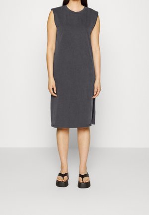 Robe sans manches gris foncé longueur genou avec fente latérale, portée avec des sandales compensées noires sur une personne debout devant un fond uni.