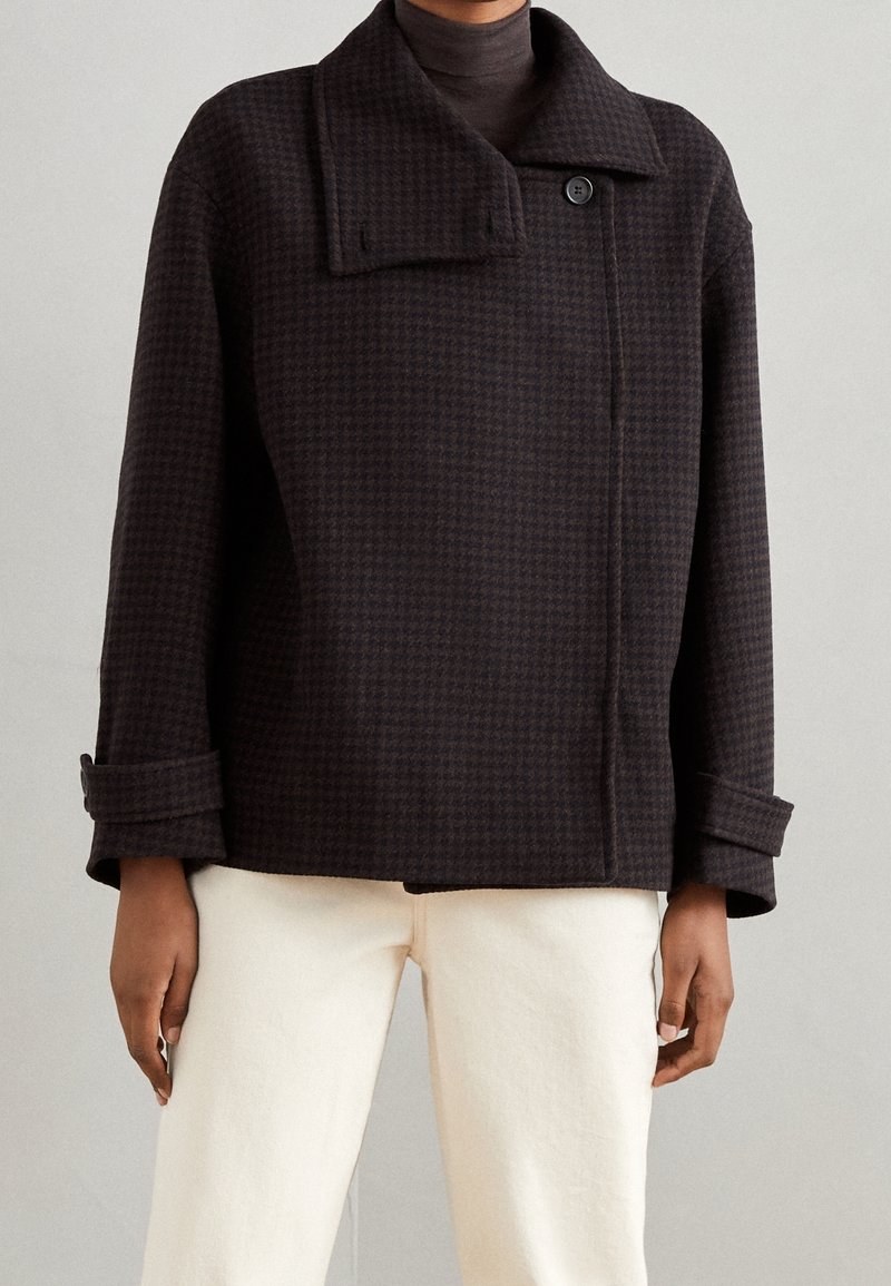 Personne portant une veste marron foncé à motif pied-de-poule avec large col, un seul bouton et manches retroussées sur un pantalon crème.