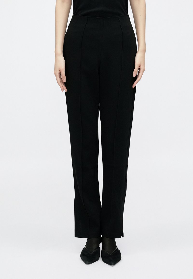 Fekete, egyenes szabású, oldalsó hasításokkal ellátott nadrág, magas derékkal és sima anyagstrukturával. Fekete, hegyes orrú cipővel viselve.