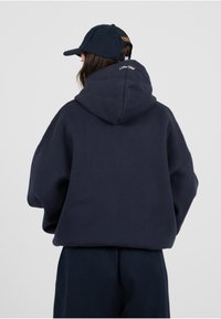 Sudadera azul marino de gran tamaño con una textura suave y una capucha grande, que presenta un pequeño bordado de logo en la parte posterior. Combinada con una gorra a juego.