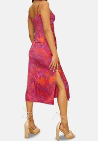 Nainen pukeutunut hihattomaan kukalliseen midimekkoon, jossa on sivuhalkio, ja vaaleanbeigeihin nilkkaremmeillä varustettuihin korkokenkiin, seisoo sivuttain.