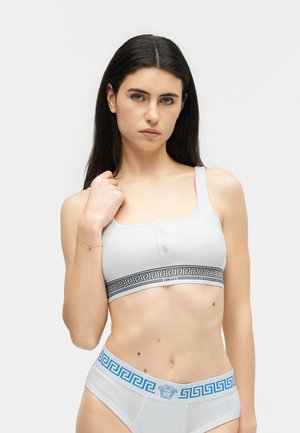 Ung kvinde med langt mørkt hår iført hvid Versace sports-bh og matchende trusser med græske nøglemønstre, stående mod en ensfarvet baggrund.