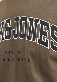 Brun bomulds t-shirt med stort hvidt og sort tekst "K&JONES" og mindre sort tekst "EST. 90 WORLD WIDE" på ryggen. Enkel design.