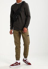 Graues Langarmshirt mit weißem Kragen, khakifarbene Jogginghose mit seitlicher Reißverschlusstasche und rote Vans-Sneaker mit weißen Schnürsenkeln.