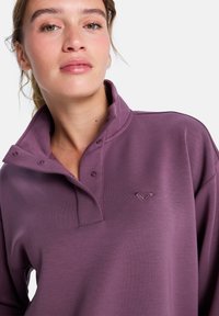 Fioletowa, długorękawowa koszulka polo z wysokim kołnierzem, zapięciami na napy oraz haftowanym logo po lewej stronie klatki piersiowej. Gładka tekstura, casualowy design.