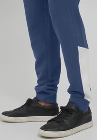 Blue sweatpants with white side panel and cuffed hems paired with black sneakers featuring a smooth texture and white sole.