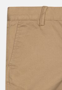 Beige chinos med en glat tekstur, der har sidelommer og et fladt frontdesign. Syede detaljer er synlige langs sømmene.