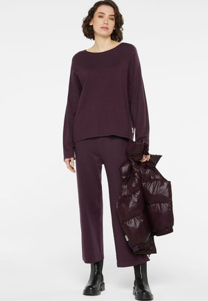 Lila, übergroßer Pullover mit weitem Ausschnitt, kombiniert mit passenden, weiten Hosen und einer glänzenden, puffigen kastanienbraunen Weste, die in einer Hand gehalten wird.