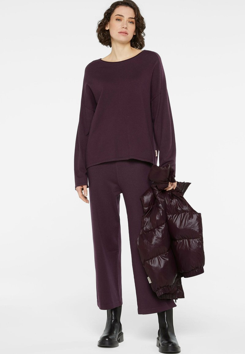 Lila, übergroßer Pullover mit weitem Ausschnitt, kombiniert mit passenden, weiten Hosen und einer glänzenden, puffigen kastanienbraunen Weste, die in einer Hand gehalten wird.