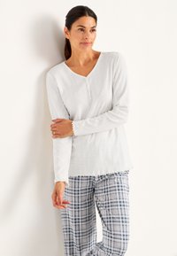 Vit långärmad topp med subtil ribbad textur, v-ringning och scalloped nederkant, tillsammans med grå rutiga loungebyxor med blå detaljer.