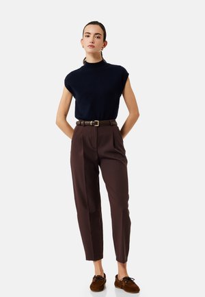 Giovane donna in piedi con le mani dietro la schiena, che indossa un top nero senza maniche, pantaloni marroni a vita alta con cintura e mocassini marroni.