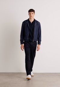 Navy zip-up jacket over a black polo, paired with black trousers and white sneakers. Smooth fabric with a relaxed fit and minimal design.