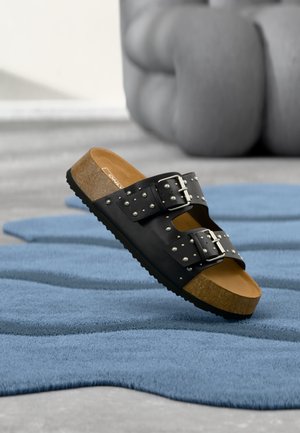 Sandalia tipo slide de cuero negro con suela de corcho, dos hebillas y tachuelas de metal, colocada sobre una superficie texturizada azul en interiores.