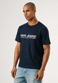 Marineblauwe katoenen t-shirt met ronde hals en korte mouwen, met witte logo-tekst en Union Jack-detail op de voorkant.