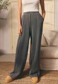 Donna che indossa pantaloni grigi scuri larghi e mocassini beige, in piedi all'interno accanto a un divano beige e a una porta bianca chiusa.