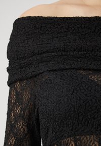 Top nero in pizzo senza spalle che presenta intricati motivi floreali, tessuto strutturato e un design aderente che arriva fino a metà braccio.