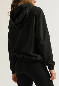 Schwarzer Kapuzenpullover mit lockerer Passform, ausgestattet mit gerippten Bündchen und Saum. Der Stoff wirkt glatt und leicht strukturiert.