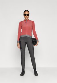 Rød langærmet cykeltrøje med lynlås og grå accenter; parret med sorte tætsiddende leggings og sorte cykelsko. Mørke solbriller bæres.