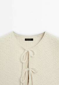Gros plan d'un cardigan en maille crème avec un col rond et deux nœuds noués à l'ouverture devant.