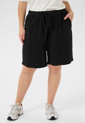 Person wearing black knee-length shorts with front pockets, white sneakers, and a white top, standing against a plain background.