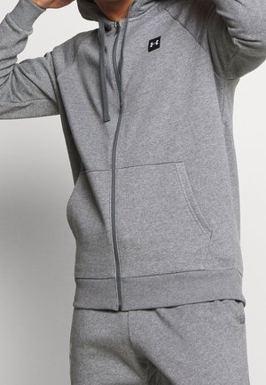 Personne portant un sweat à capuche gris Under Armour avec fermeture éclair et un pantalon de survêtement assorti, ajustant la capuche avec les mains près de la tête.