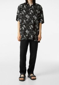 Camisa negra de manga corta con patrones gráficos blancos que incluyen animales y símbolos. Combinada con pantalones cargo negros y sandalias oscuras.