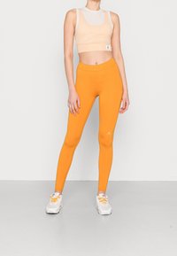 Persikofärgad kort topp med meshdetaljer och orangea leggings, båda i elastisk tyg. Sneakers är vita med orange snörning. Enkel design.