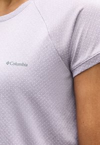 Κοντινό πλάνο ενός ατόμου που φοράει ανοιχτό μοβ κοντομάνικο πουκάμισο με διακριτικό σχέδιο και λογότυπο Columbia στο στήθος.