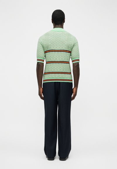 Muž stojaci chrbtom, oblečený v svetlozelenom pletenom tričku s krátkym rukávom a oranžovými, čiernymi a bielymi pruhmi a tmavých nohaviciach s úzkymi pruhmi.