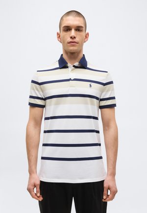 Uomo che indossa una polo a maniche corte con righe orizzontali blu navy e beige, colletto blu navy e piccolo logo sul petto sinistro, in piedi davanti a uno sfondo semplice.