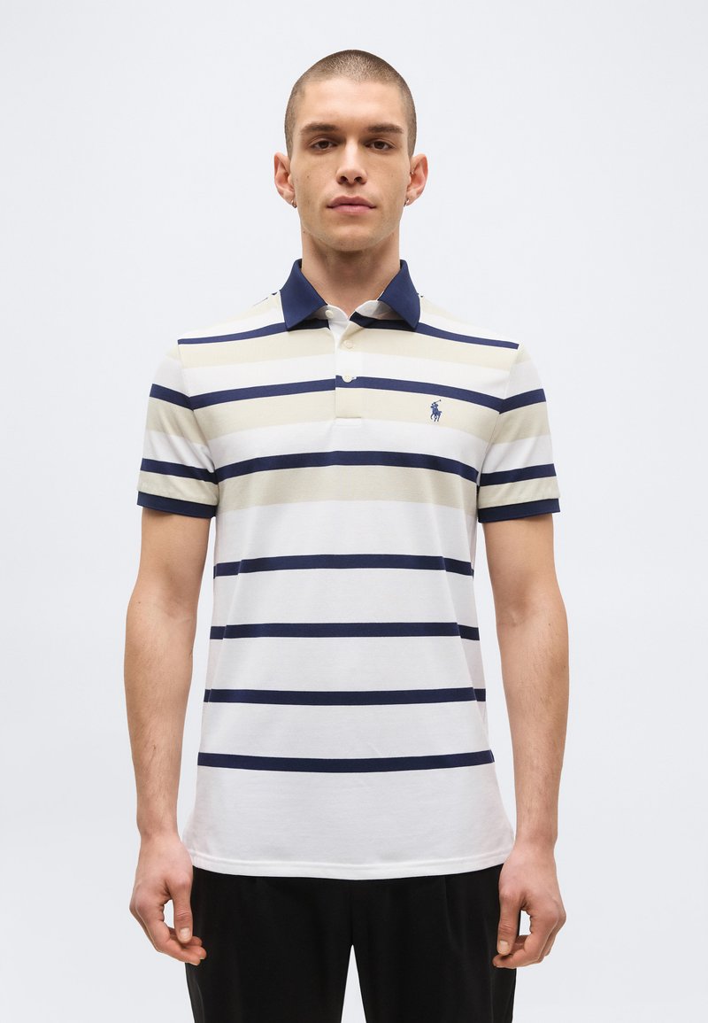 Homme portant un polo manches courtes avec des rayures horizontales bleu marine et beige, col bleu marine et petit logo sur la poitrine gauche, debout devant un fond uni.
