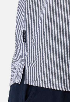 Camicia a righe caratterizzata da strisce verticali blu e bianche alternate, con un’apertura laterale e un'etichetta "SEASIDE" sull'orlo. Tessuto in cotone.