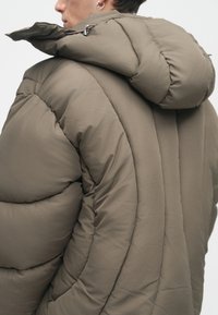 Casaco puffer em verde-azeitona com gola alta, apresentando painéis acolchoados e um capuz ajustável, com detalhes como um brilho subtil e fechos de correr elegantes.