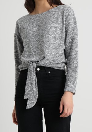 Femme portant un haut en maille gris à manches longues avec un détail noué à l'avant, associé à un pantalon noir taille haute.
